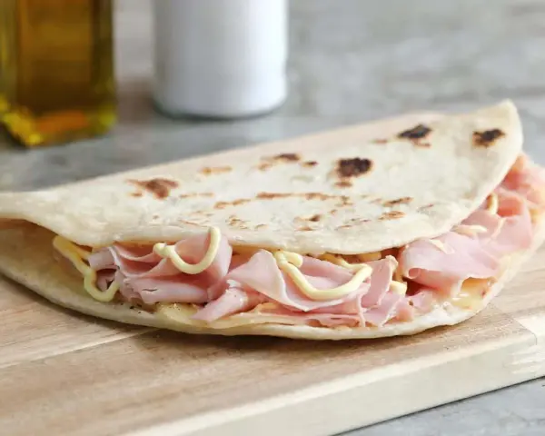 Piadina sfogliata romagnola ripiena di prosciutto cotto e formaggio, su tagliere di legno.