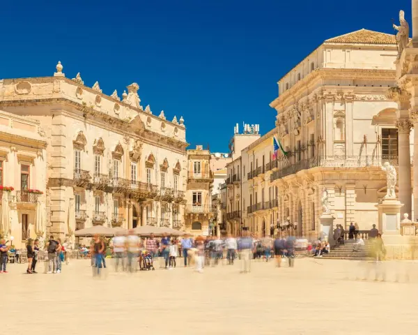 Piazza affollata con edifici storici. La gente si muove rapidamente, quasi sfocata, mentre si valuta la **classificazione stelle hotel** per un soggiorno in questa splendida cornice.