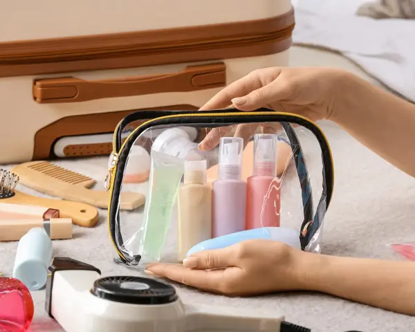 Mani che preparano cosa mettere nel beauty case: flaconi, gel e accessori da viaggio in una trousse trasparente.