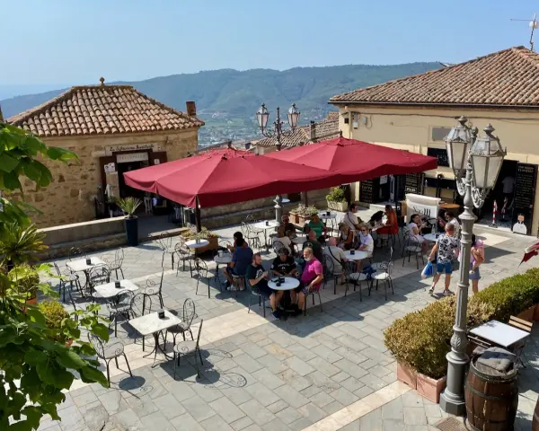 Piazza soleggiata a Castellabate con tavolini all'aperto e gente che si gode il momento. La scritta "qui non si muore" invita a rilassarsi.