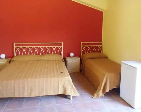 Camera semplice in un albergo una stella: due letti singoli e un letto matrimoniale con copriletti color ocra, pareti rosse e gialle, comodini e un piccolo frigorifero.