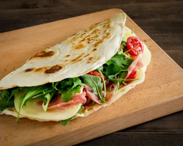Piadina ricetta: focaccia ripiena di prosciutto, formaggio, rucola e pomodorini su tagliere di legno.