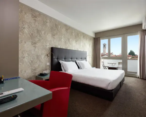 Camera moderna con letto matrimoniale, scrivania in vetro, poltrona rossa e balcone con vista. I prezzi su Booking sono a persona.