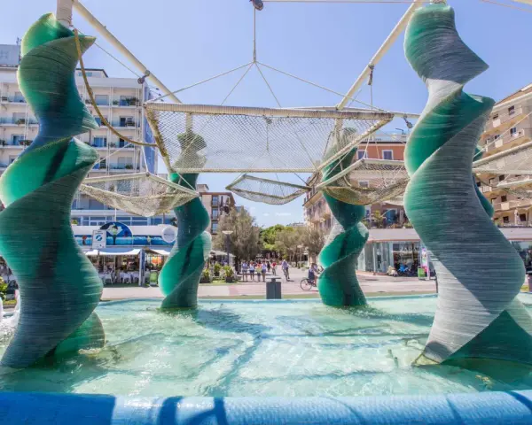 Sculture a spirale in vetro verde in una fontana a Riccione, con edifici e persone sullo sfondo.