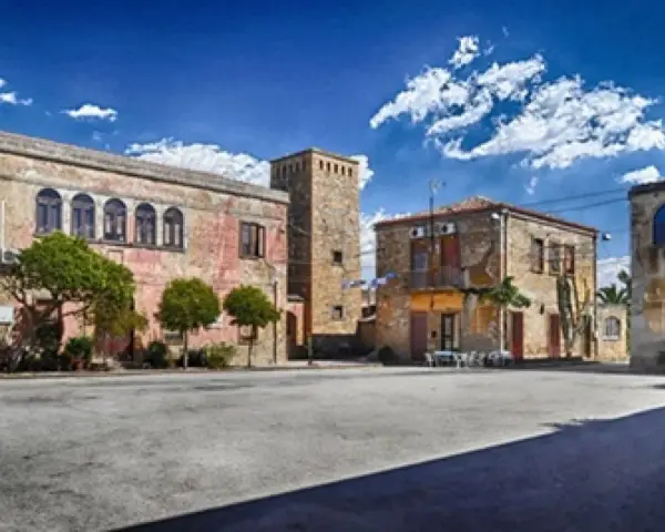 Piazza con palazzi fascisti, un torrione in pietra e alberi verdi sotto un cielo azzurro con nuvole bianche.