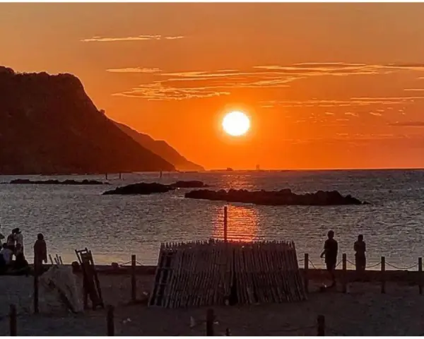 Un magico tramonto a Pesaro, con il sole che infuoca il mare e le colline sullo sfondo. Persone godono della spiaggia.