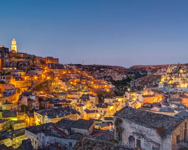Un viaggio on the road in Basilicata, con Matera illuminata al crepuscolo. Case antiche e chiese rupestri brillano di luci calde.
