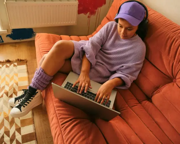 Ragazza con cuffie e cappellino viola lavora al laptop su un divano arancione. Si può andare in hotel a 16 anni, ma lei preferisce casa.