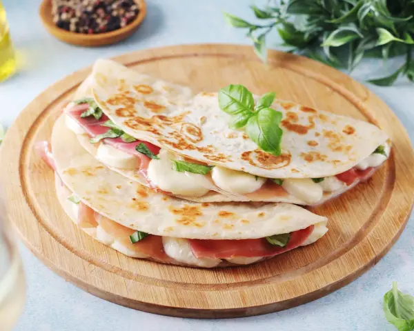 Piadina romagnola ripiena di prosciutto crudo, mozzarella e basilico, servita su un tagliere di legno.
