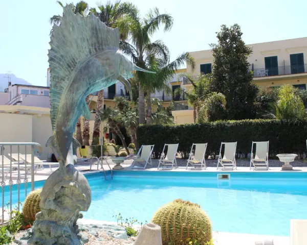 Scultura di pesce e piscina invitante al **hotel Punta del Sole Ischia**, circondata da palme e lettini.