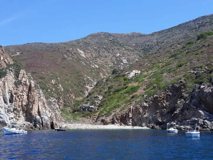 Barche a vela nella cala del corvo, con scogliere rocciose e vegetazione mediterranea.