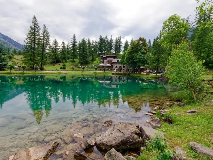 Lago alpino con riflessi verdi, perfetto per le gite in Piemonte. Case e alberi incorniciano la riva.