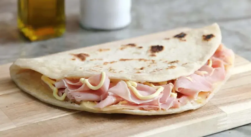 Piadina sfogliata romagnola ripiena di prosciutto cotto e formaggio, su tagliere di legno.