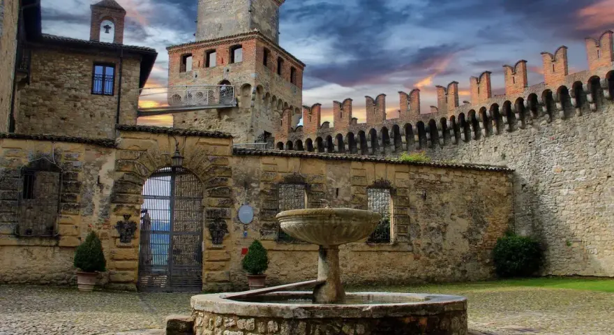 Cortile di uno dei castelli vicino Parma, con fontana in pietra e mura merlate sotto un cielo drammatico.