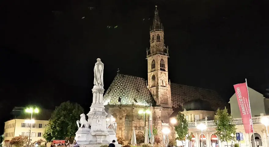 Piazza Walther di Bolzano di notte: la cattedrale, il monumento e la bandiera rossa. Un'idea di cosa vedere a Bolzano in un giorno.