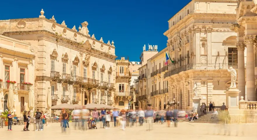 Piazza affollata con edifici storici. La gente si muove rapidamente, quasi sfocata, mentre si valuta la **classificazione stelle hotel** per un soggiorno in questa splendida cornice.