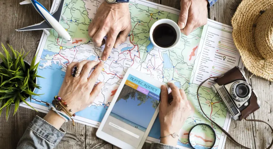 Pianificare viaggi senza agenzia: mani che studiano una mappa, un tablet con sito di viaggi, aereo giocattolo e macchina fotografica vintage.