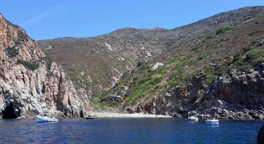 Barche a vela nella cala del corvo, con scogliere rocciose e vegetazione mediterranea.