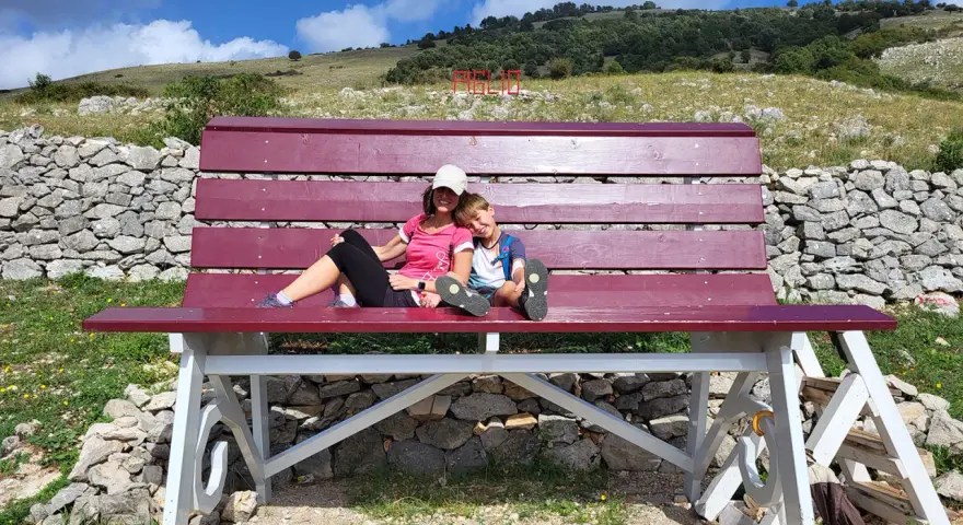 Gita domenicale con bambini nel Lazio: mamma e figlio si godono il panorama su una panchina gigante, con montagne e cielo azzurro sullo sfondo.