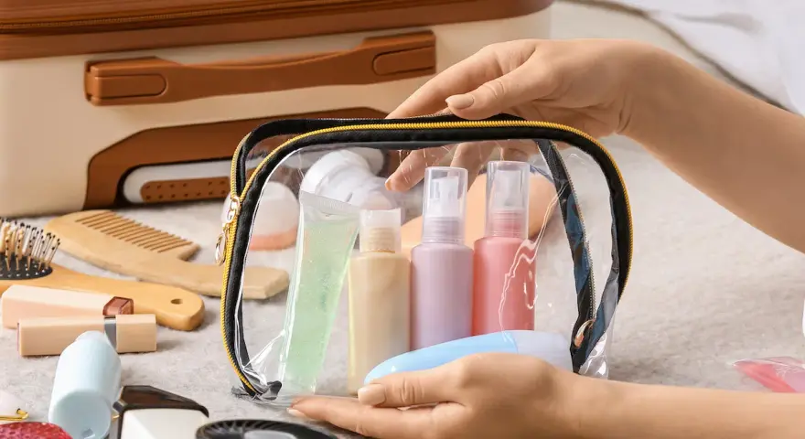 Mani che preparano cosa mettere nel beauty case: flaconi, gel e accessori da viaggio in una trousse trasparente.
