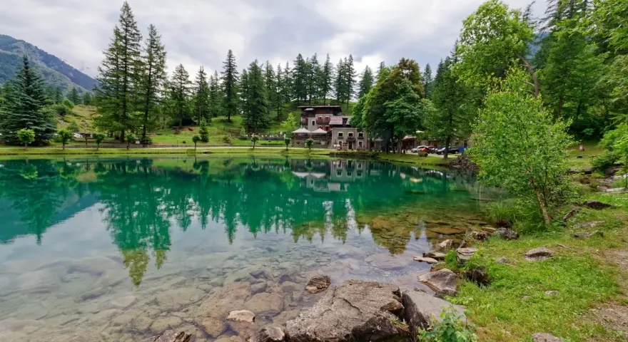 Lago alpino con riflessi verdi, perfetto per le gite in Piemonte. Case e alberi incorniciano la riva.