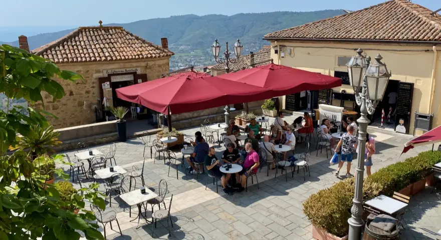 Piazza soleggiata a Castellabate con tavolini all'aperto e gente che si gode il momento. La scritta "qui non si muore" invita a rilassarsi.