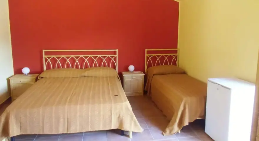 Camera semplice in un albergo una stella: due letti singoli e un letto matrimoniale con copriletti color ocra, pareti rosse e gialle, comodini e un piccolo frigorifero.