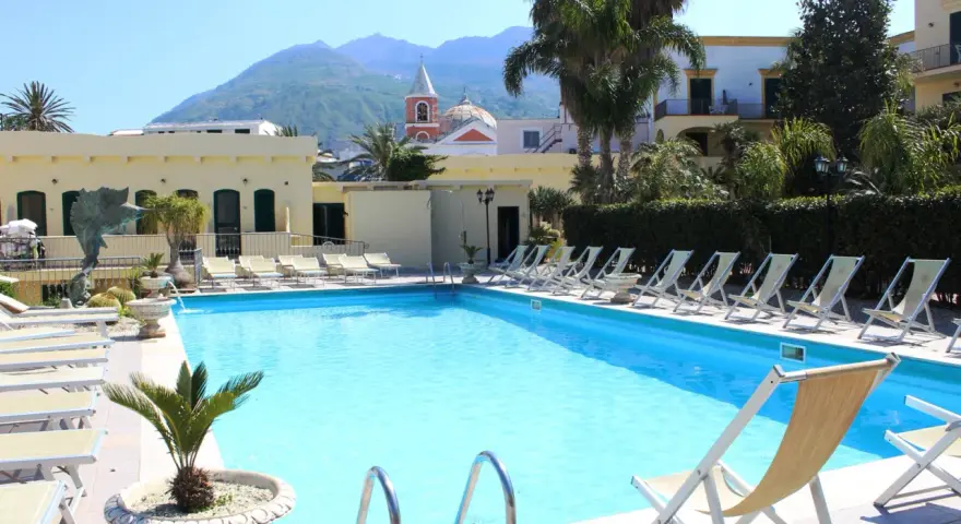 Piscina con lettini e palme, vista su montagne e chiesa. Un luogo ideale per rilassarsi, come suggeriscono le hotel terme punta del sole recensioni.