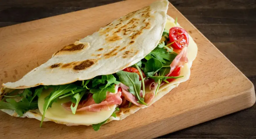 Piadina ricetta: focaccia ripiena di prosciutto, formaggio, rucola e pomodorini su tagliere di legno.