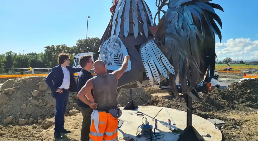 Operai al lavoro su un imponente gallo romagnolo in metallo, un'opera d'arte che domina il paesaggio.