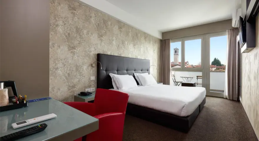 Camera moderna con letto matrimoniale, scrivania in vetro, poltrona rossa e balcone con vista. I prezzi su Booking sono a persona.