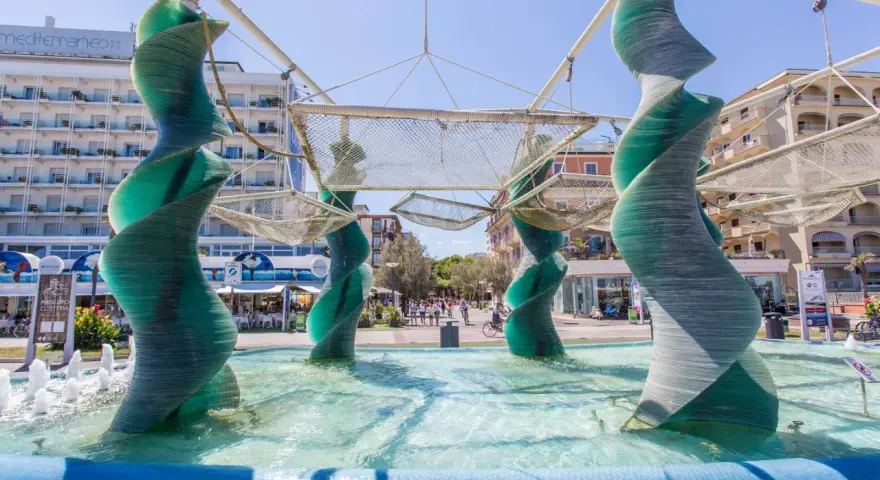 Sculture a spirale in vetro verde in una fontana a Riccione, con edifici e persone sullo sfondo.