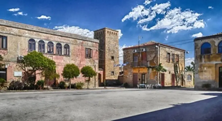 Piazza con palazzi fascisti, un torrione in pietra e alberi verdi sotto un cielo azzurro con nuvole bianche.