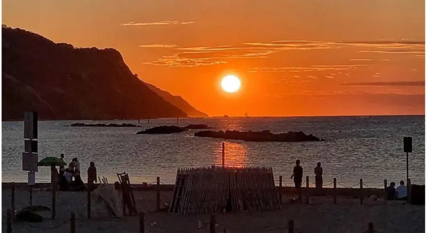 Un magico tramonto a Pesaro, con il sole che infuoca il mare e le colline sullo sfondo. Persone godono della spiaggia.