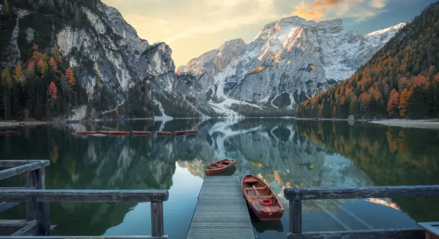 Lago di Braies, con montagne innevate e boschi autunnali riflessi sull'acqua. Per sapere come arrivare al lago di Braies in treno da Roma, cerca online.