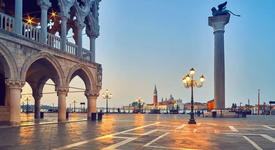 L'alba illumina il colonnato del Palazzo Ducale, proiettando lunghe ombre sulla piazza. In lontananza, il leone alato veglia su Piazza San Marco.