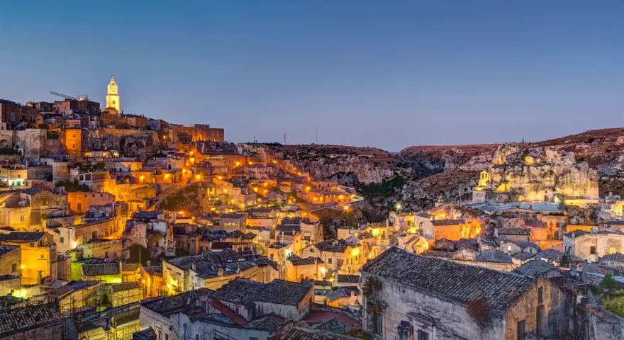 Un viaggio on the road in Basilicata, con Matera illuminata al crepuscolo. Case antiche e chiese rupestri brillano di luci calde.