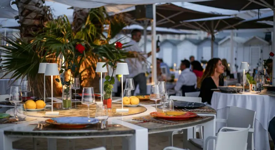 Tavoli apparecchiati sulla spiaggia di Riccione, con limoni, rose rosse e piccole lampade. Atmosfera serale, perfetta per una cena di manna.