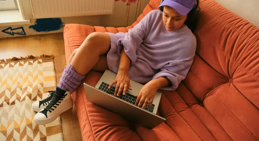Ragazza con cuffie e cappellino viola lavora al laptop su un divano arancione. Si può andare in hotel a 16 anni, ma lei preferisce casa.