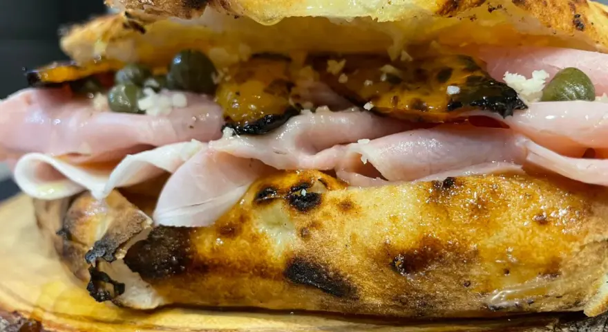 Un panino gourmet con mortadella, capperi e pomodori secchi, un vero re artù a Lecce, servito su un tagliere di legno.