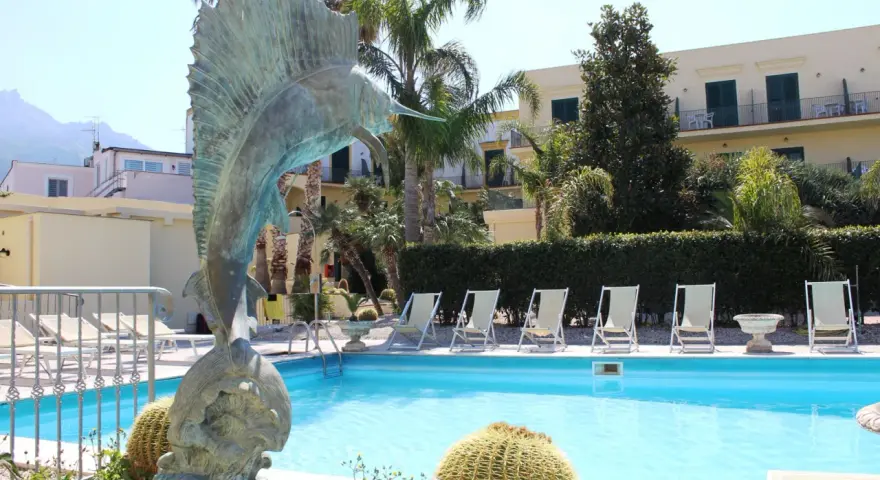 Scultura di pesce e piscina invitante al **hotel Punta del Sole Ischia**, circondata da palme e lettini.