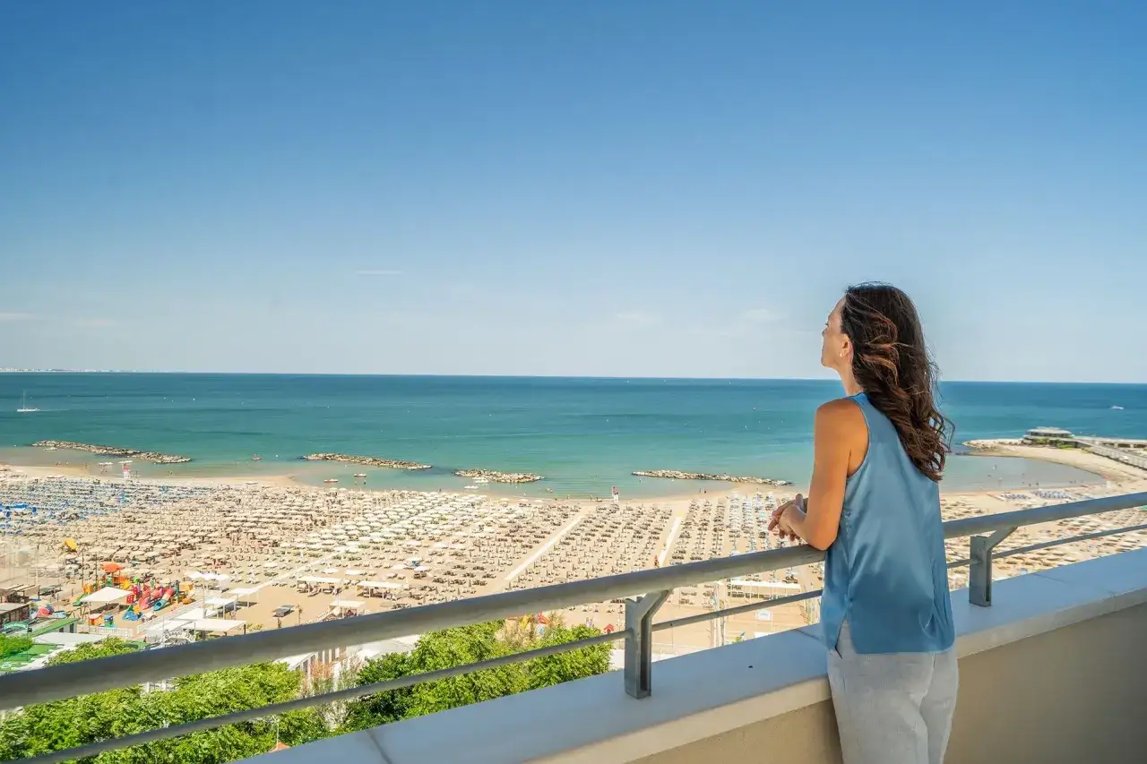 Vista panoramica su una spiaggia affollata di Cattolica, con ombrelloni e lettini che si estendono fino al mare azzurro. Una donna ammira il paesaggio dalla terrazza.