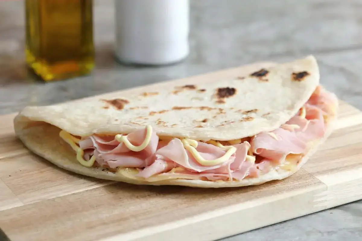 Piadina sfogliata romagnola ripiena di prosciutto e formaggio, servita su un tagliere di legno.