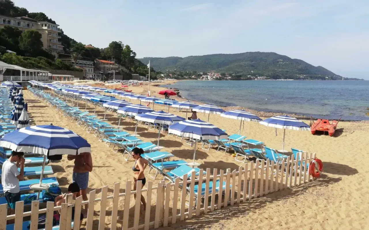 Un posto al mare a Castellabate: file ordinate di lettini e ombrelloni blu e bianchi sulla sabbia dorata, con il mare azzurro e le colline verdi sullo sfondo.