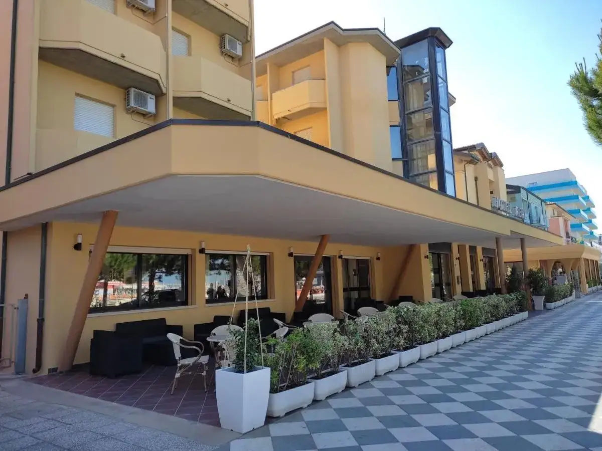 L'Hotel Real Villamarina, con la sua facciata gialla e l'ascensore in vetro, offre un'accogliente area esterna con tavoli e divanetti, circondata da aiuole fiorite.