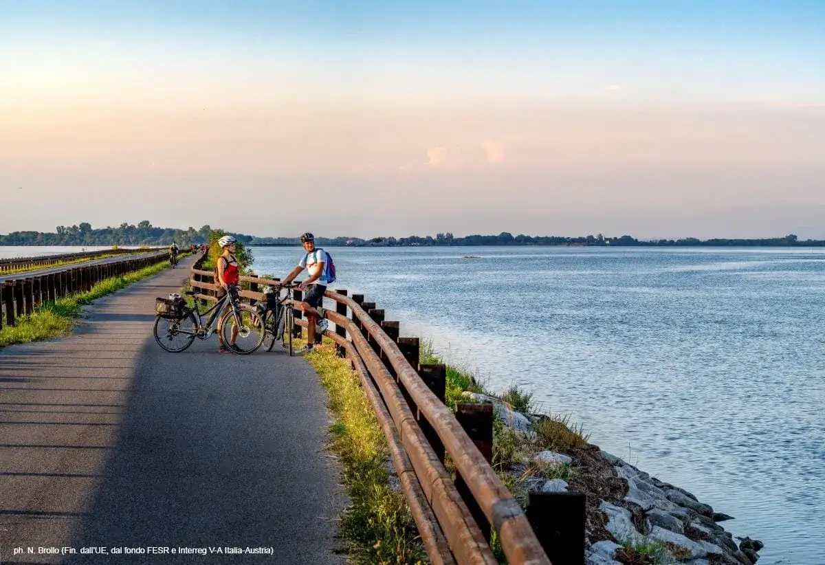 Coppia in bicicletta si gode il tramonto su una delle splendide piste ciclabili del Friuli Venezia Giulia, con il mare calmo a fianco.