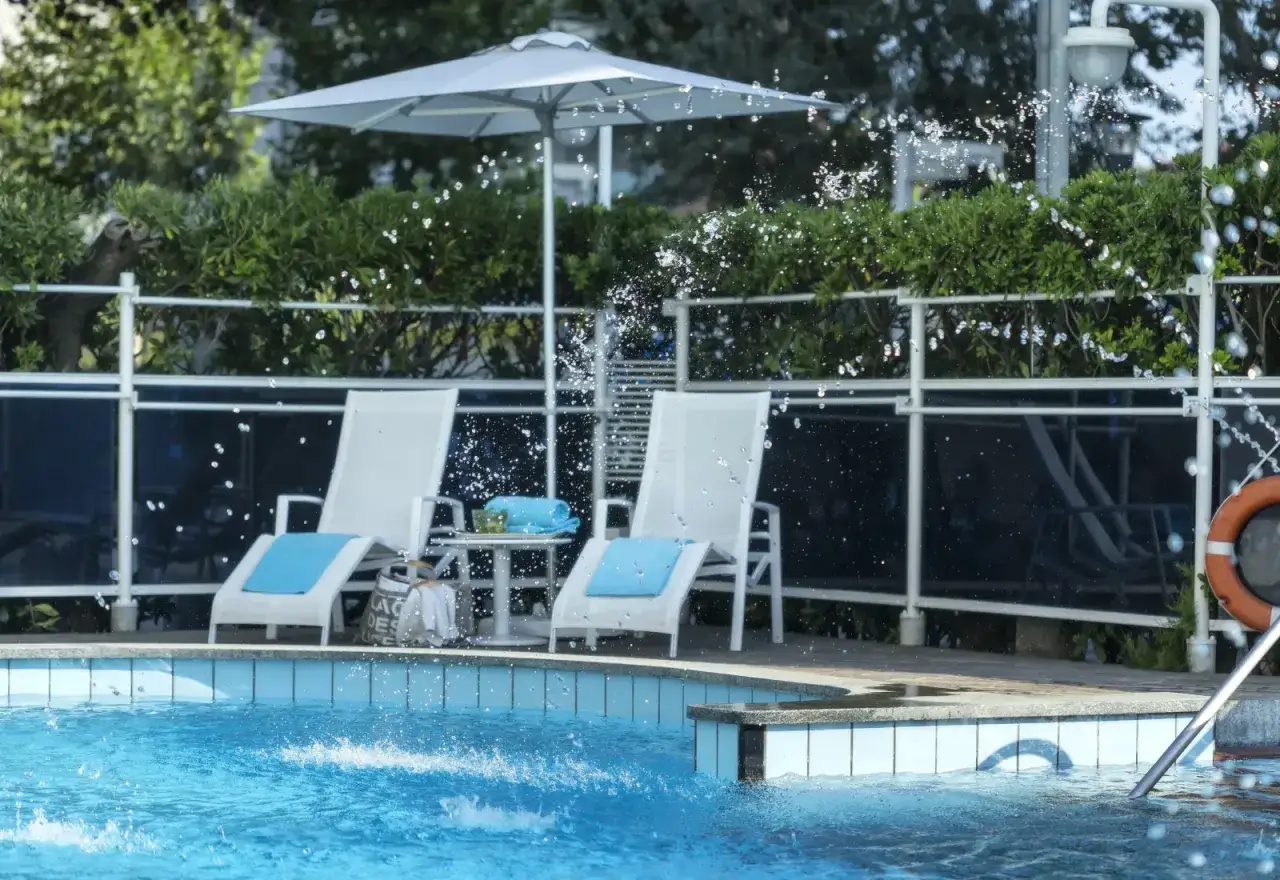 Lettini bianchi con cuscini azzurri e un ombrellone bianco vicino alla piscina del hotel Abners. L'acqua schizza vivacemente.