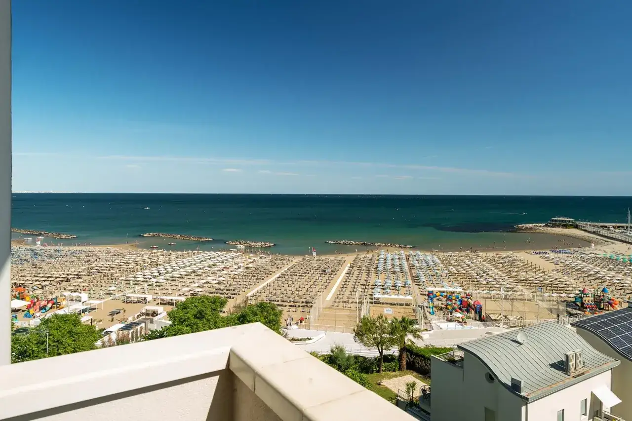 Vista dall'alto di una spiaggia affollata con ombrelloni e lettini, un mare azzurro e un cielo limpido. Sembra una piccola acropoli cattolica di relax.
