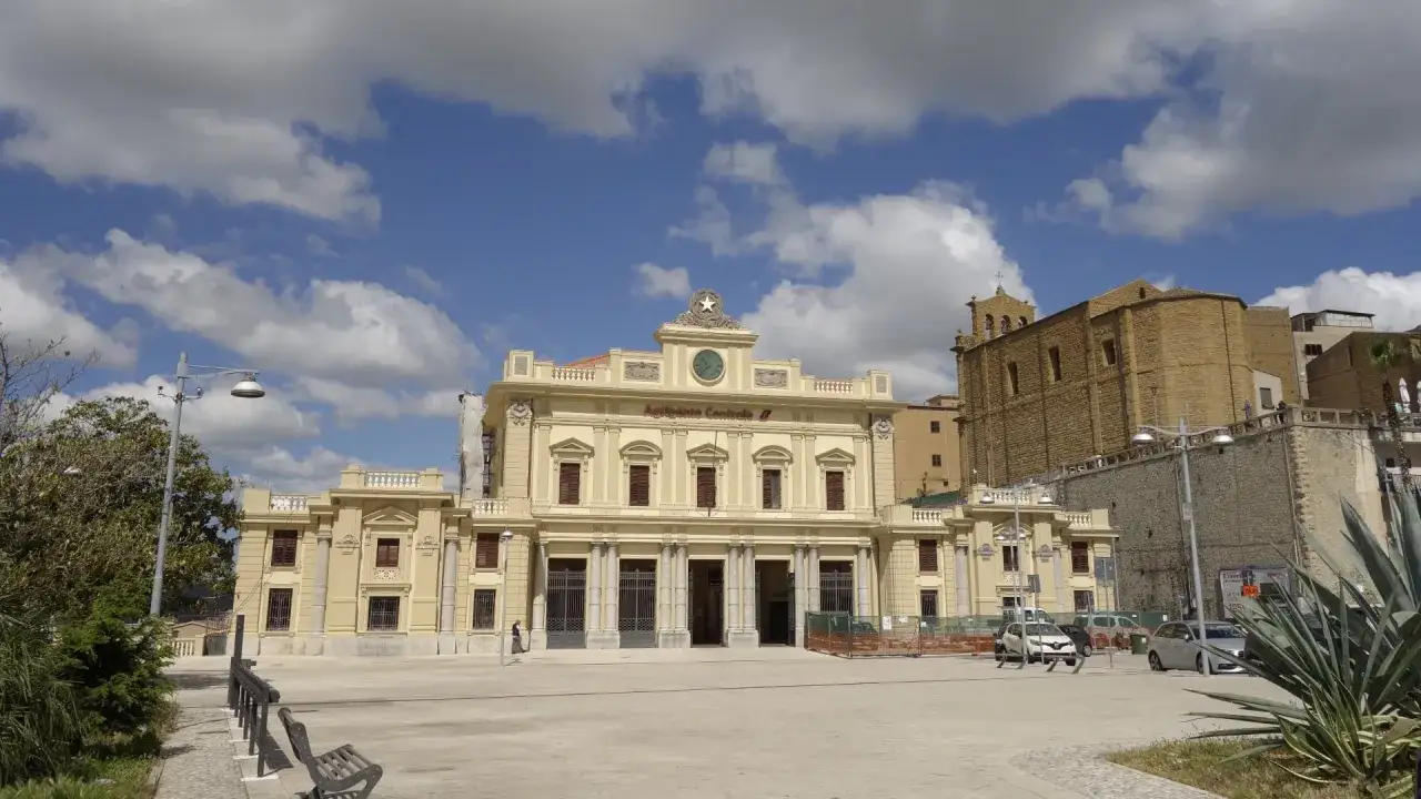 Stazione centrale di Agrigento, un esempio di palazzi fascisti con la sua imponente facciata gialla e il cielo azzurro con nuvole bianche.