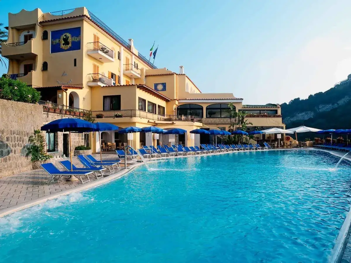 Piscina e lettini al hotel terme tramonto d'oro Ischia, con l'edificio giallo sullo sfondo e il cielo azzurro.
